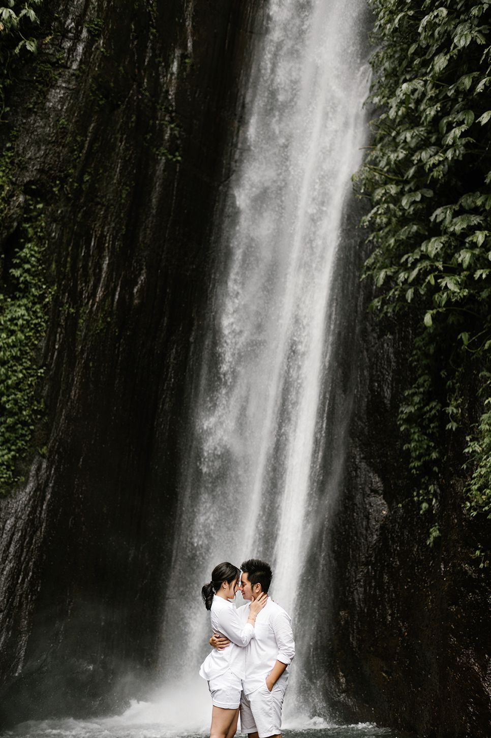 Couple in white outfits sharing a romantic kiss in front of a stunning tropical waterfall surrounded by lush greenery.