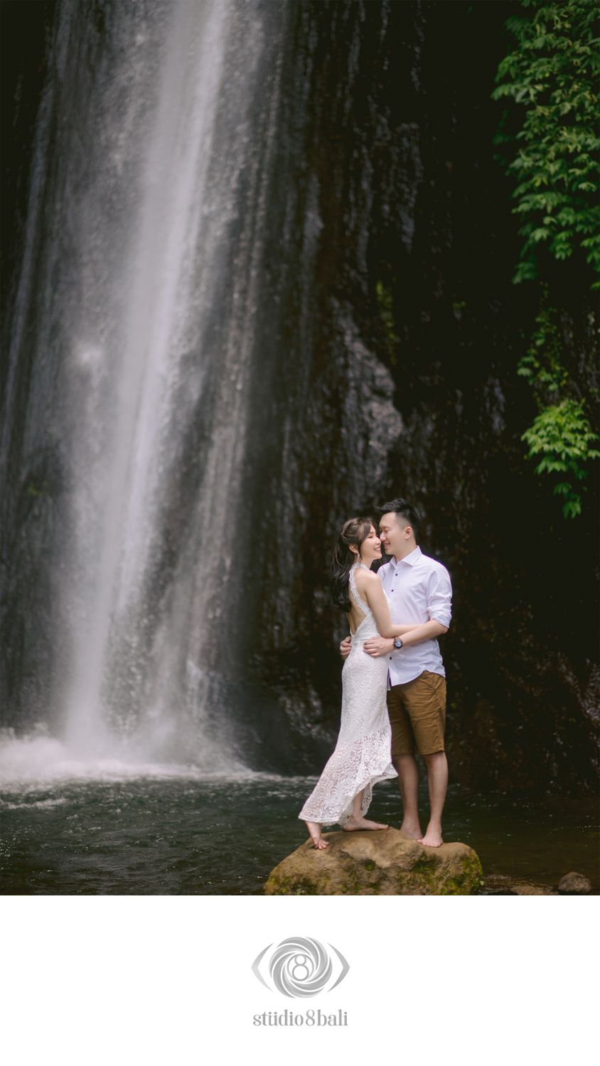 Couple embracing romantically in front of a stunning waterfall in Bali, wearing white dress and casual attire.