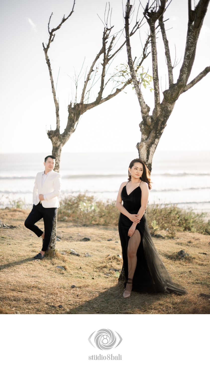 Couple poses near bare trees by the ocean, woman in black gown, man in white suit, beach pre-wedding shoot.