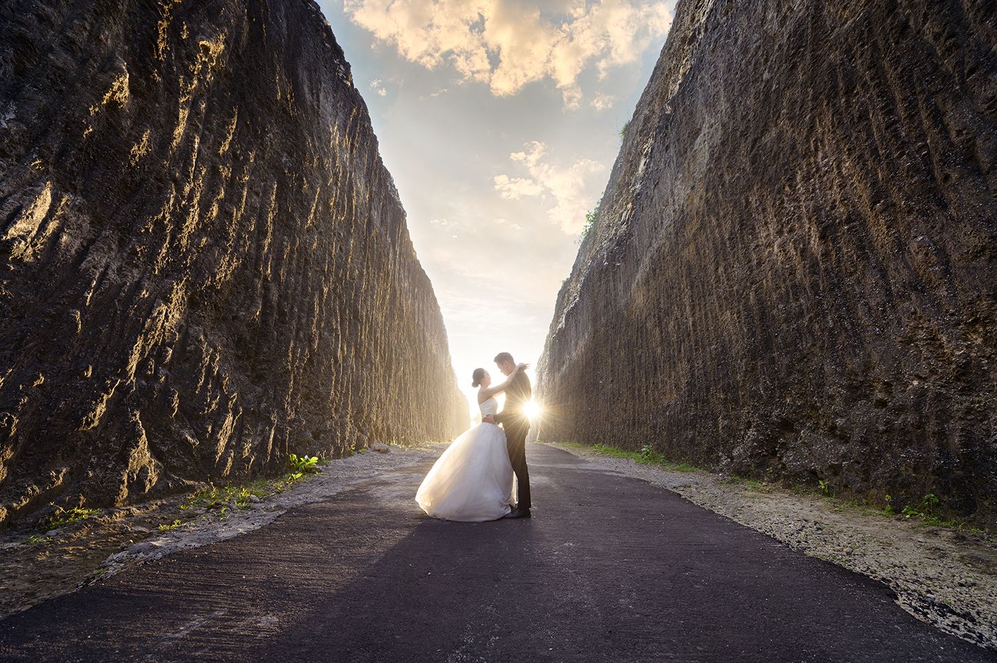Bride and groom embracing on a road between towering rock walls at sunset, golden light glowing behind them.