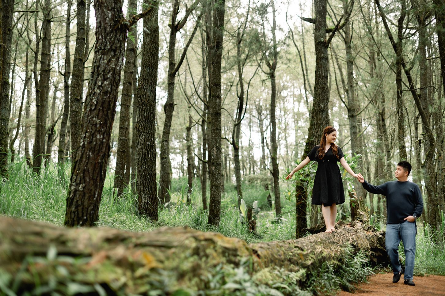 Couple sharing a romantic moment in a lush pine forest, woman in black dress standing on fallen log.