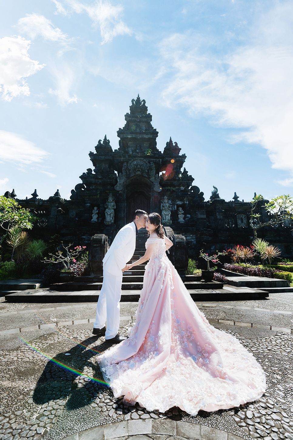 Couple in wedding attire kissing before ornate Balinese temple, bride in pink floral gown with long train.