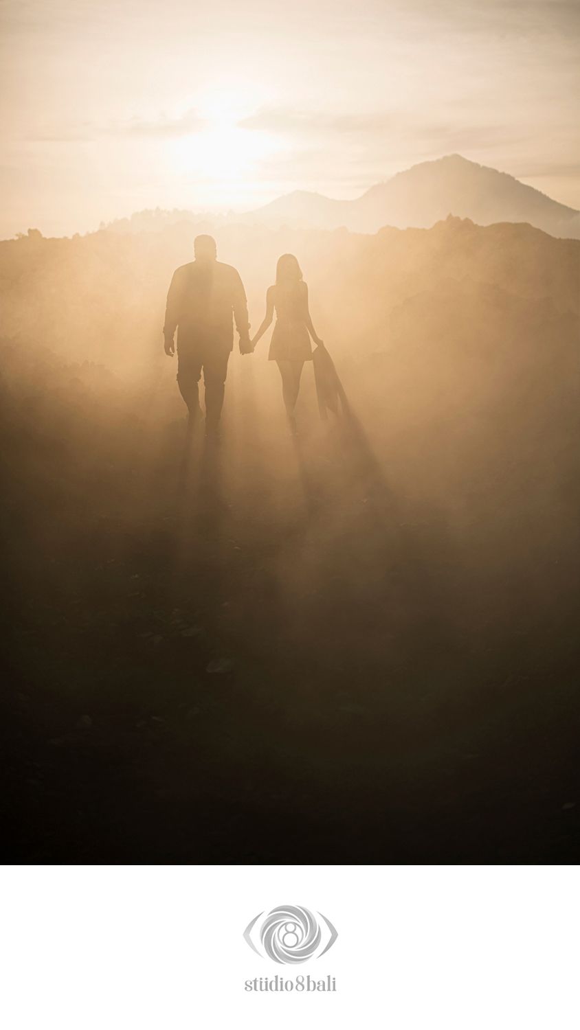 Couple holding hands walking toward misty golden sunset with mountain silhouettes in romantic backlit landscape photo.