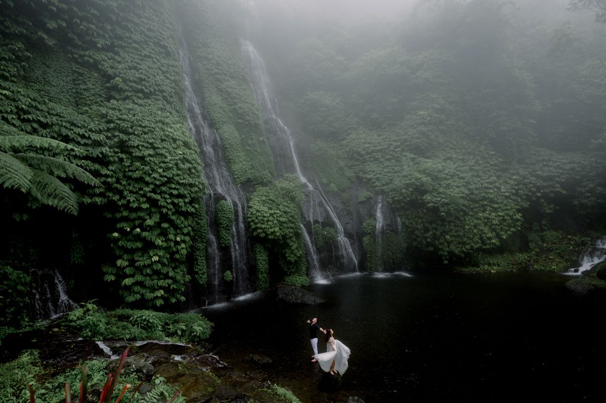 Misty waterfall cascades over green cliffs as a couple in white embraces by a serene jungle pool.