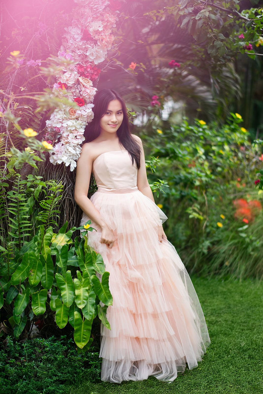 Young woman in a strapless blush pink tiered tulle gown posing beside a floral tree in a garden.