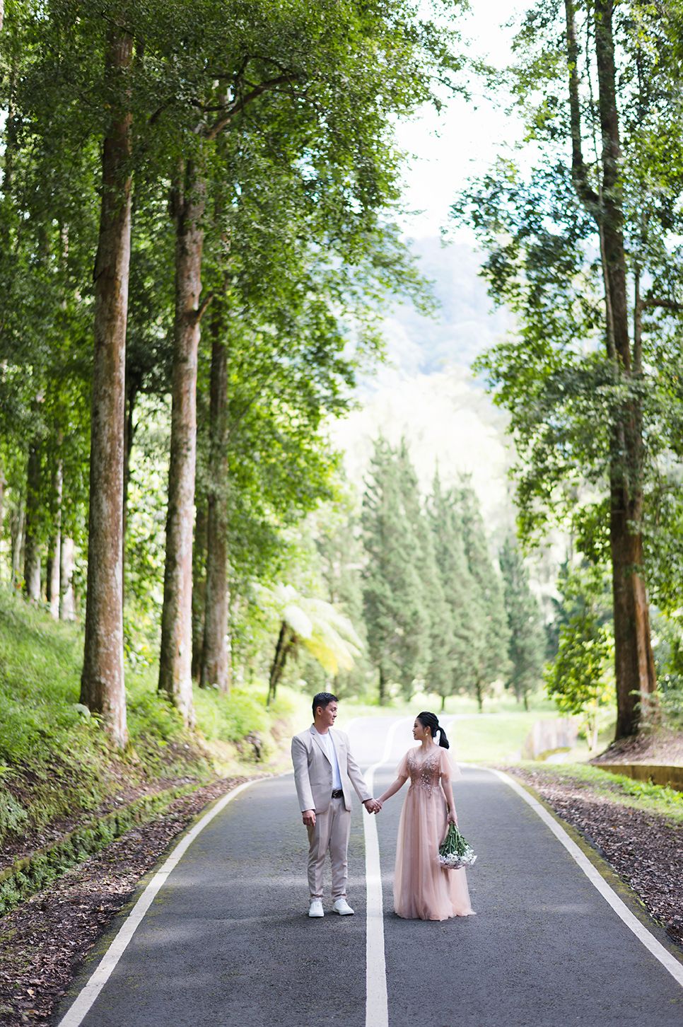 Couple in wedding attire holding hands on a tree-lined road surrounded by lush green forest.
