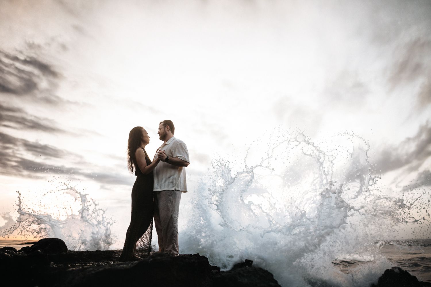 Couple embraces on rocky shore as dramatic ocean wave crashes behind them at sunset.