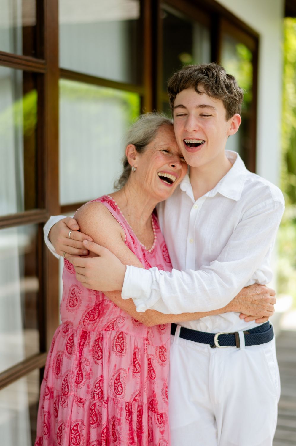A laughing teenage boy in white shirt hugs a smiling older woman in pink dress outdoors.