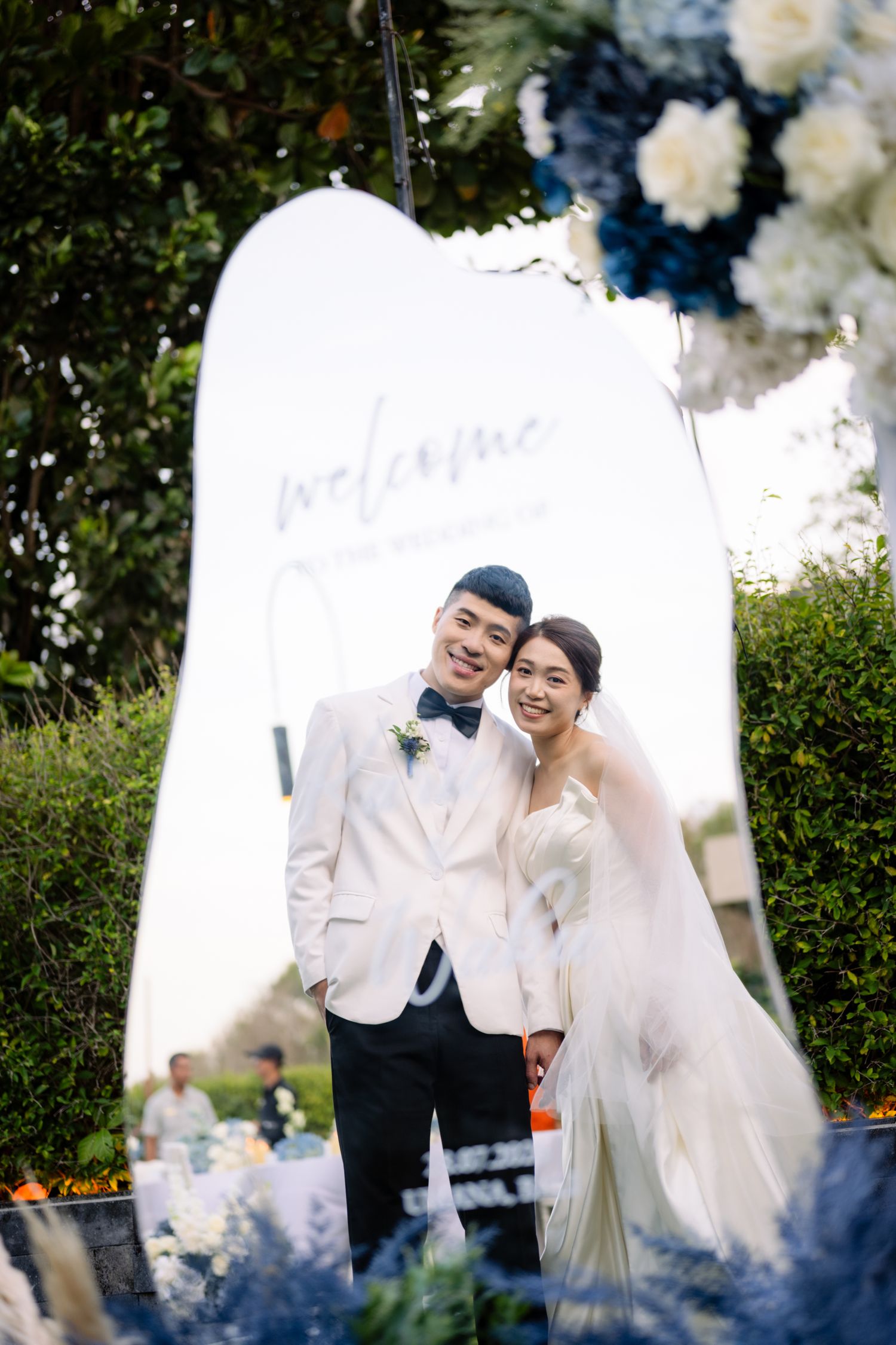 Smiling bride in a white gown and groom in a white tuxedo jacket pose beside a welcome sign at an outdoor garden wedding with blue flowers.