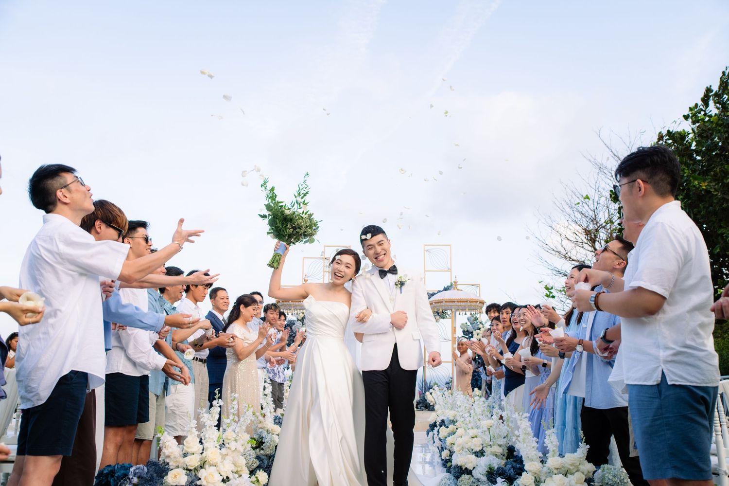 Bride in a white gown and groom in a white suit walk down a floral aisle as joyful wedding guests cheer and toss petals outdoors.