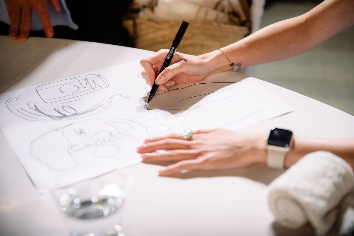Woman wearing a smart watch draws a sketch with a black marker on white paper on a table beside a rolled towel and a glass.