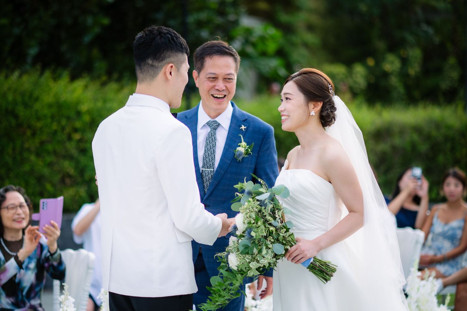 Smiling officiant in a blue suit leads an outdoor wedding ceremony as the bride in a white veil holds a green bouquet and faces the groom.