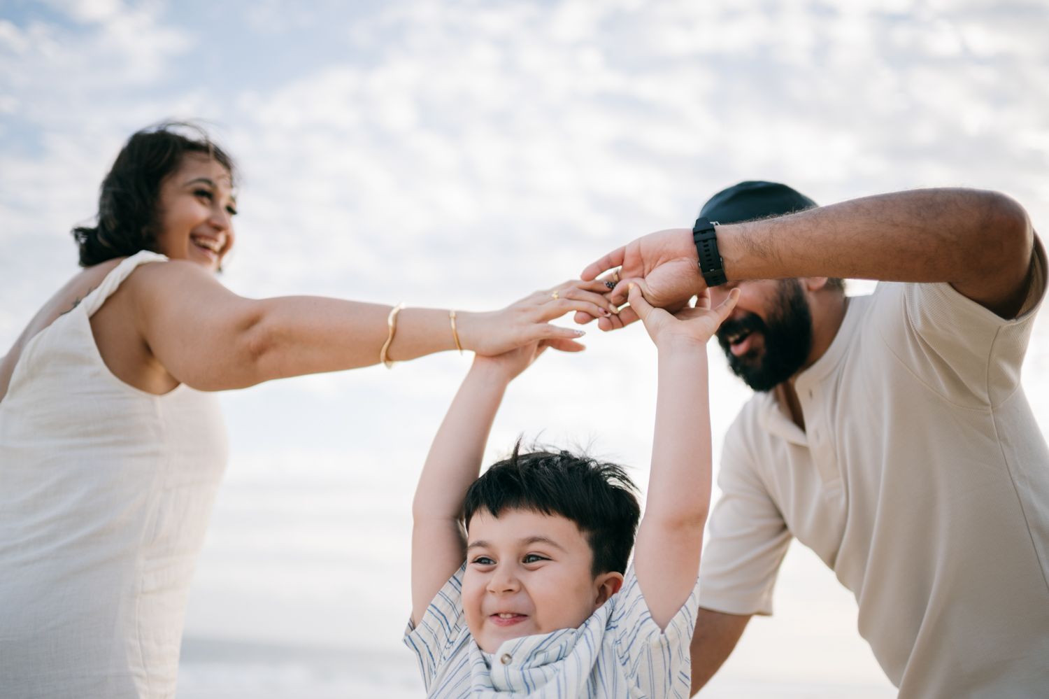 Happy family with parents lifting young child on beach, smiling together against cloudy sky backdrop.