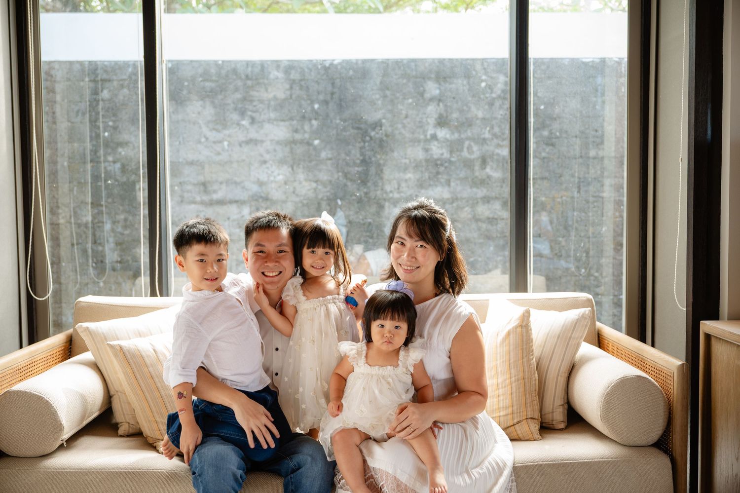 Happy Asian family of five smiling together on a white sofa by large windows in a bright modern living room.