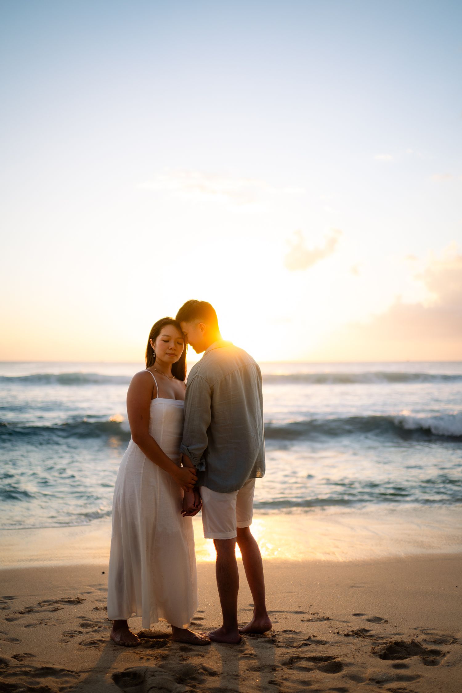 Couple holding hands on a sandy beach at sunset, woman in white dress, man in gray blazer, ocean waves behind them.
