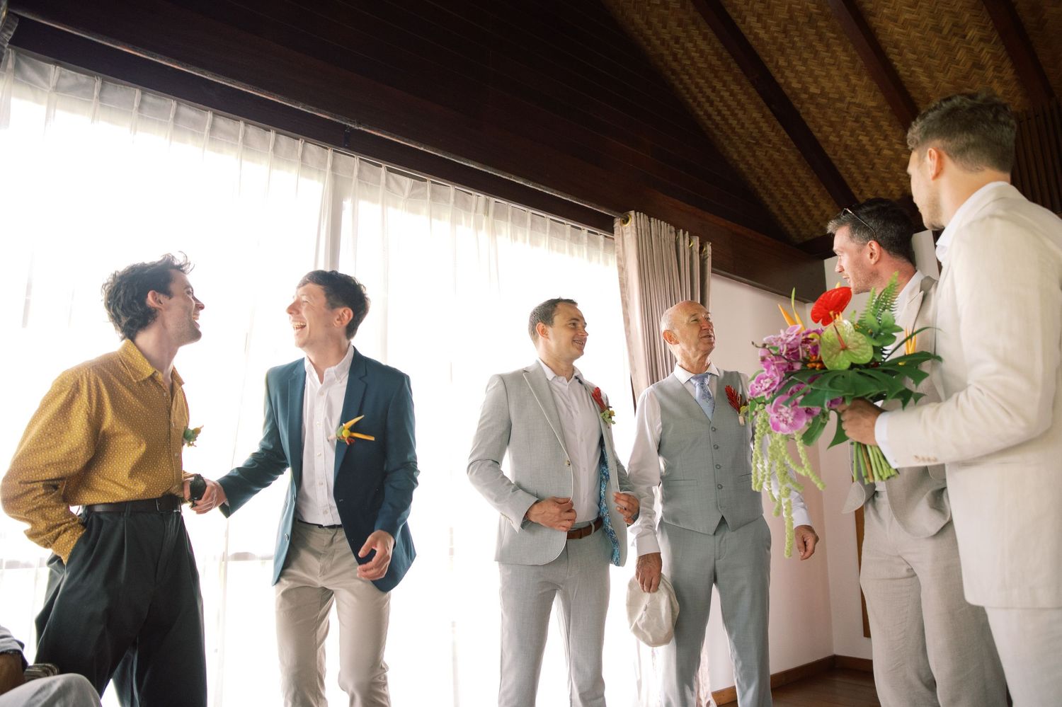 Groomsmen in suits shaking hands and holding flowers before a wedding ceremony indoors.