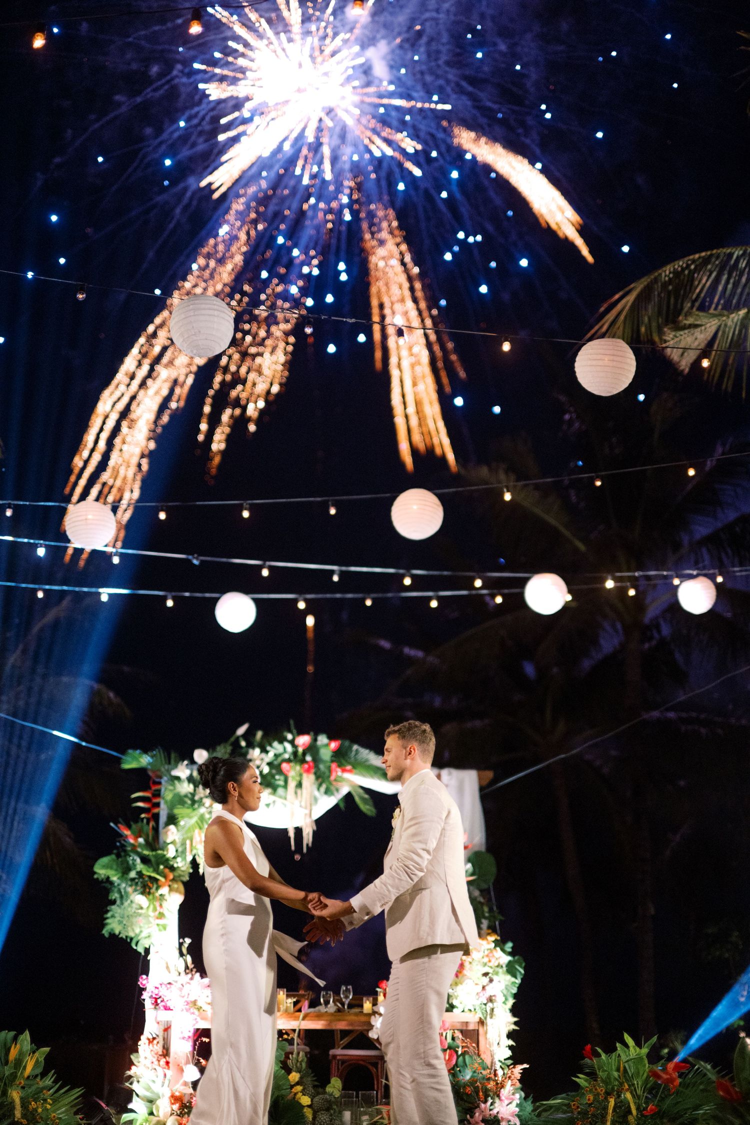 Couple holds hands at outdoor wedding ceremony with fireworks and string lights illuminating the night sky.