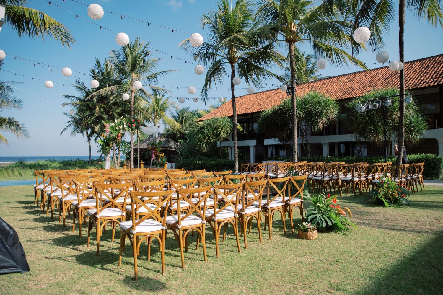Outdoor wedding ceremony setup with wooden cross-back chairs, palm trees, and white paper lantern decorations on a tropical lawn.