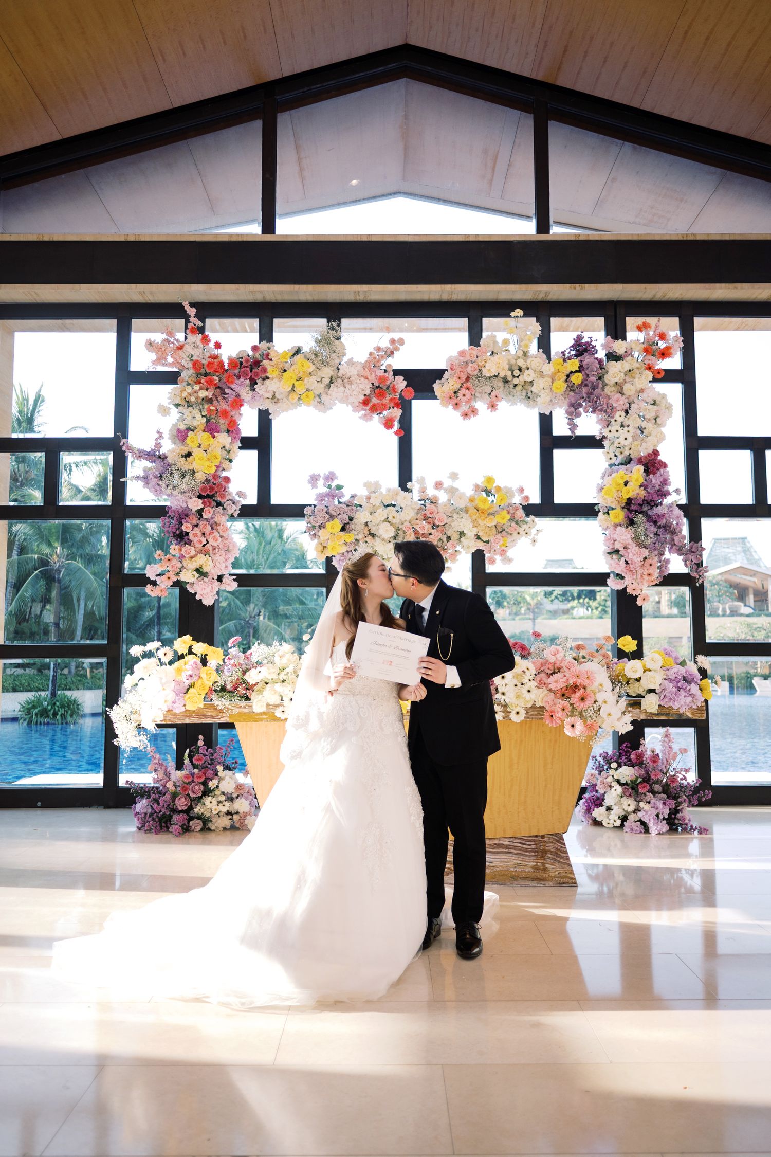 Bride and groom kissing under a colorful floral arch during their wedding ceremony indoors.