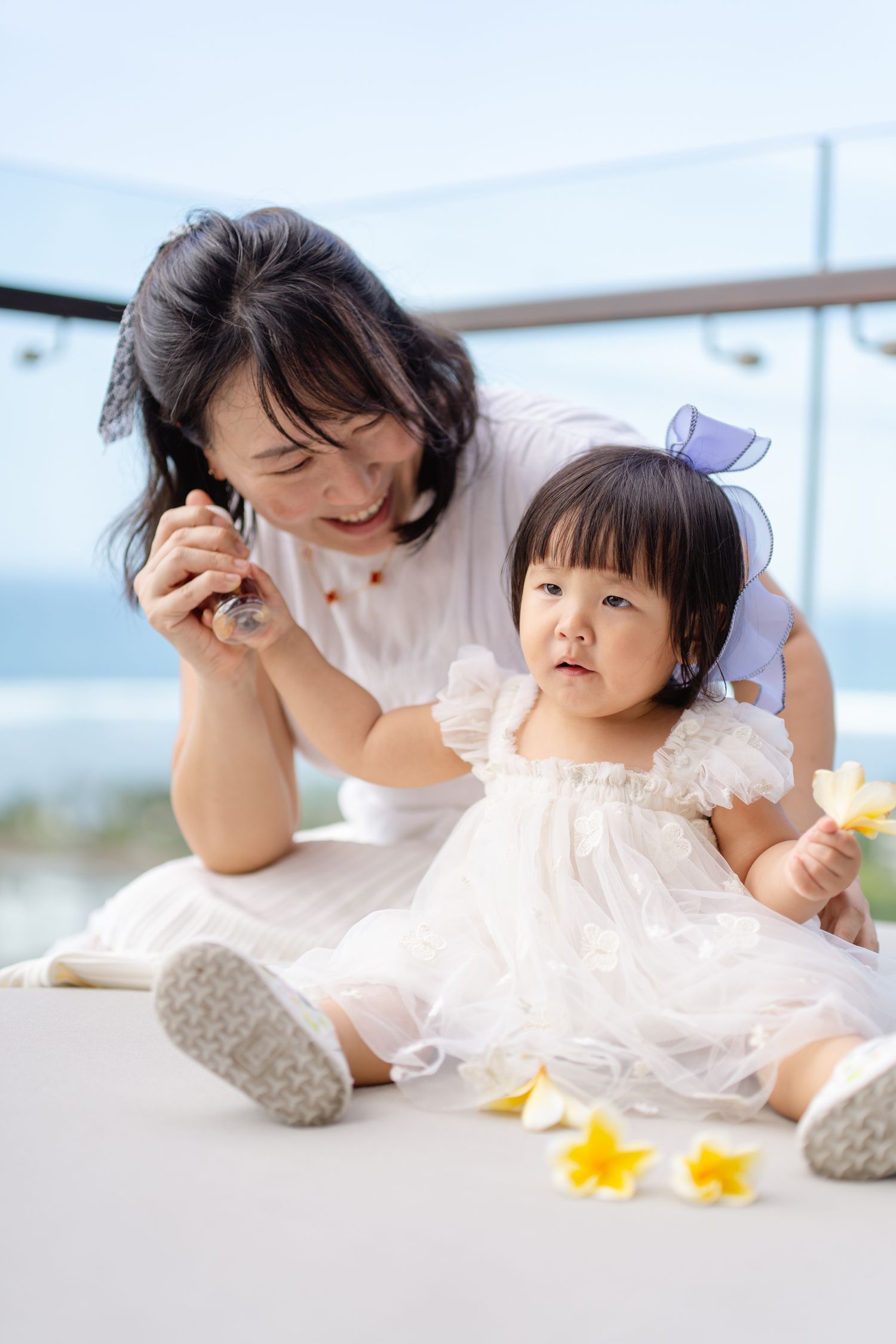 Asian mother and baby girl in white dresses sitting together on balcony with ocean view and yellow flowers.