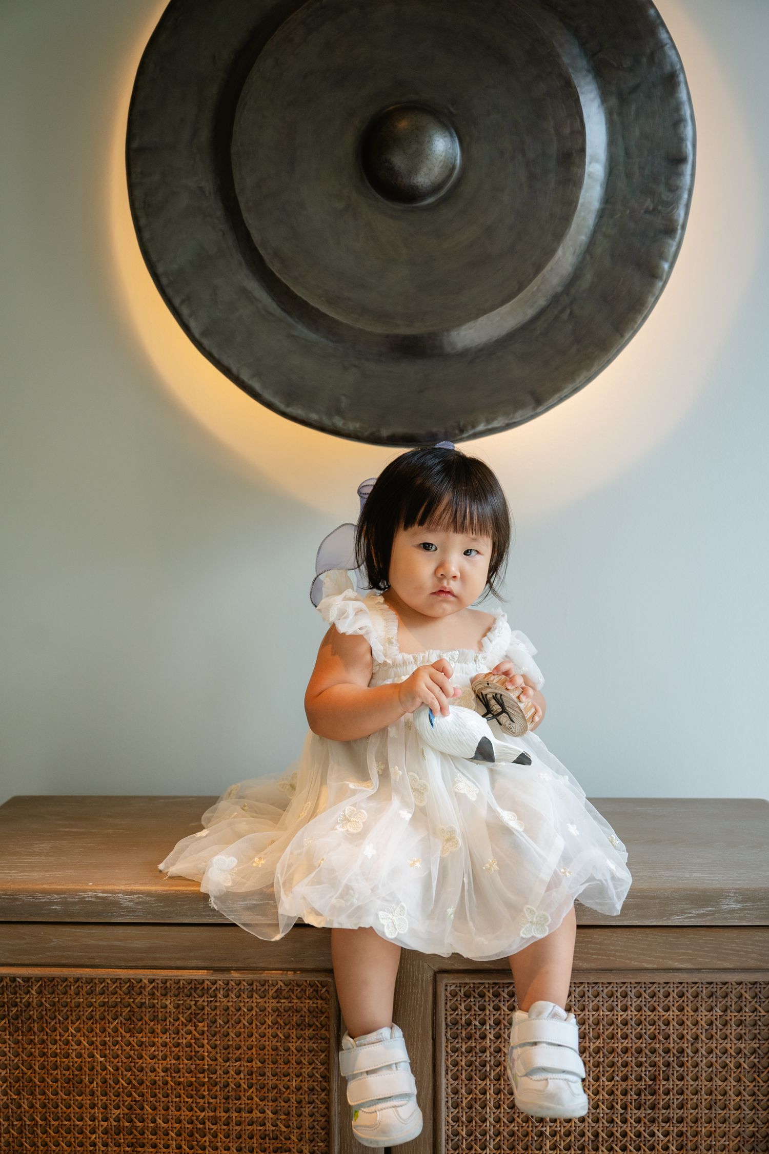 Toddler girl in white fairy dress with wings sits on wooden cabinet before large decorative gong.