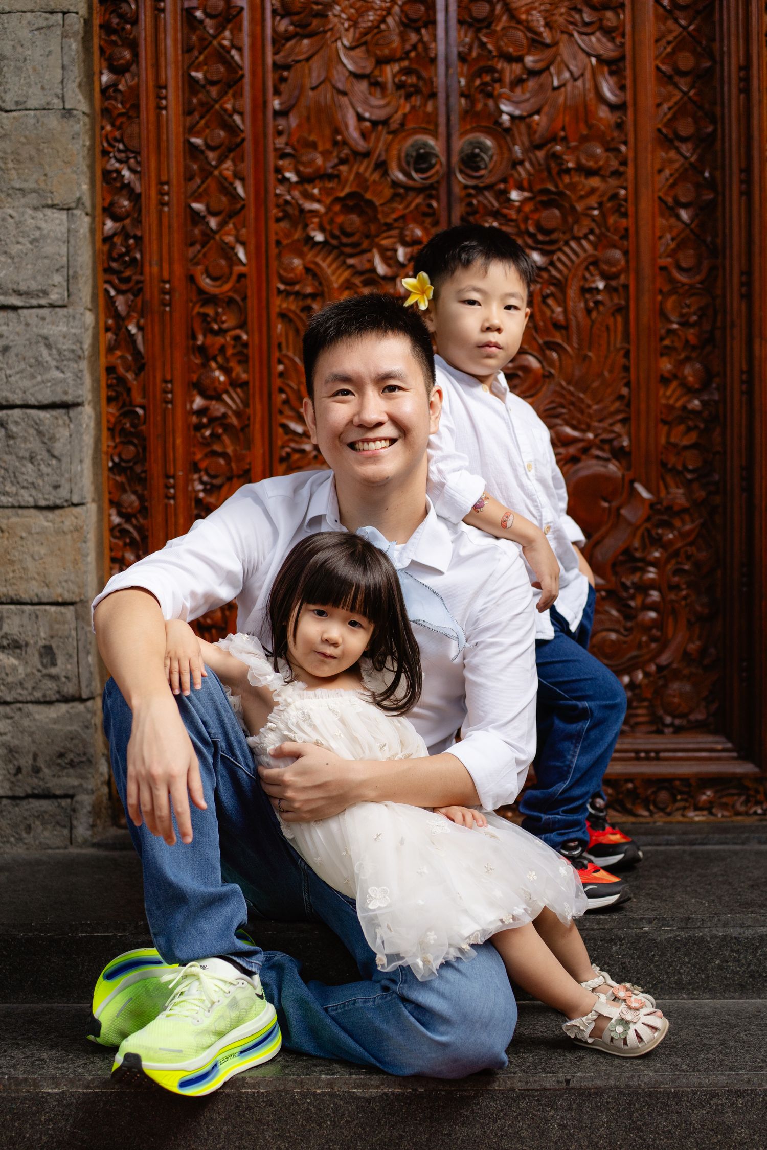 A smiling Asian man in a white shirt sits on steps with two young children in front of ornate wooden doors.