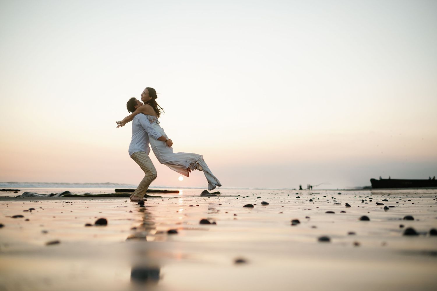 Couple embracing and spinning on a beach at sunset, wearing white outfits with ocean waves in the background.