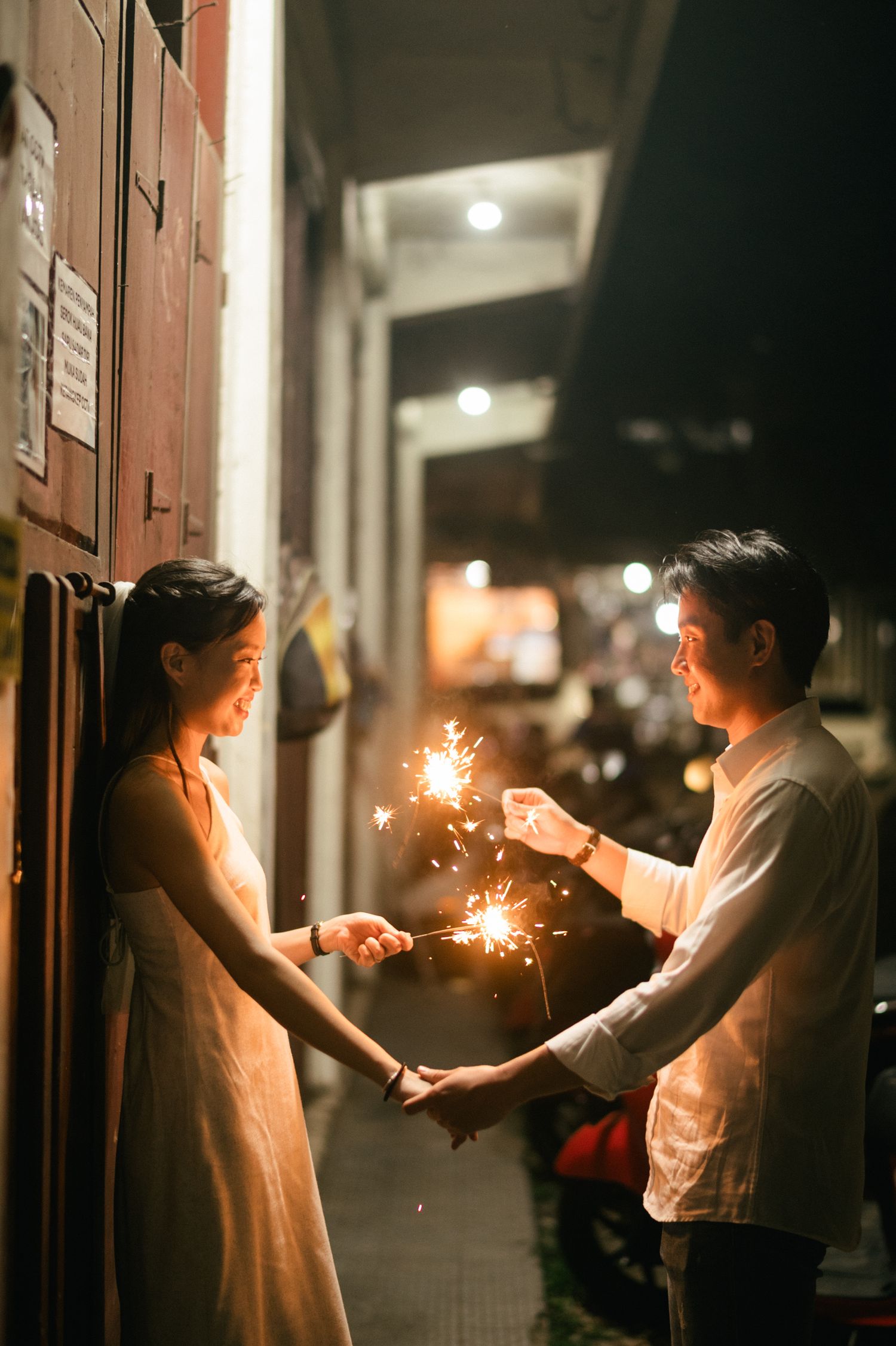Couple holding sparklers together at night in a romantic alley, woman in white dress, man in white shirt.