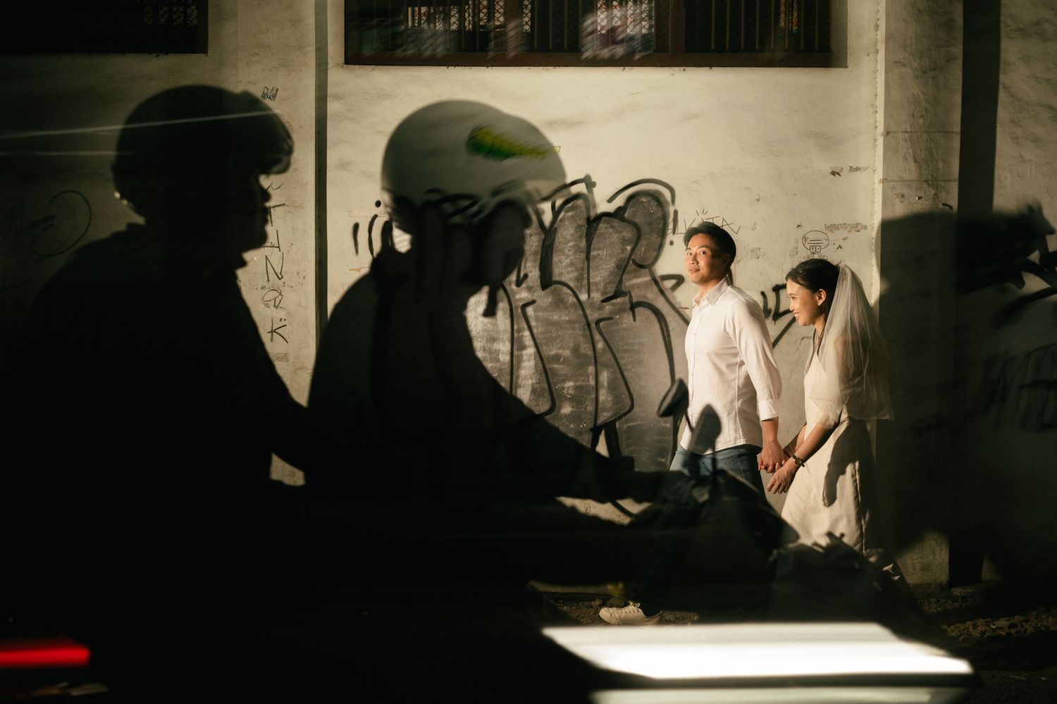 Couple in wedding attire poses near graffiti wall at night with motorcyclist silhouette in foreground.