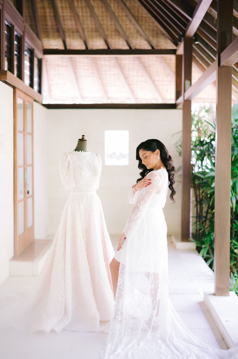 Bride in lace robe admires elegant pink wedding gown on mannequin in bright Balinese villa setting.