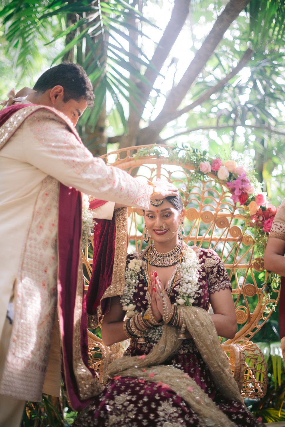 Indian bride in purple lehenga sits smiling during traditional Hindu wedding ceremony outdoors under floral arch.
