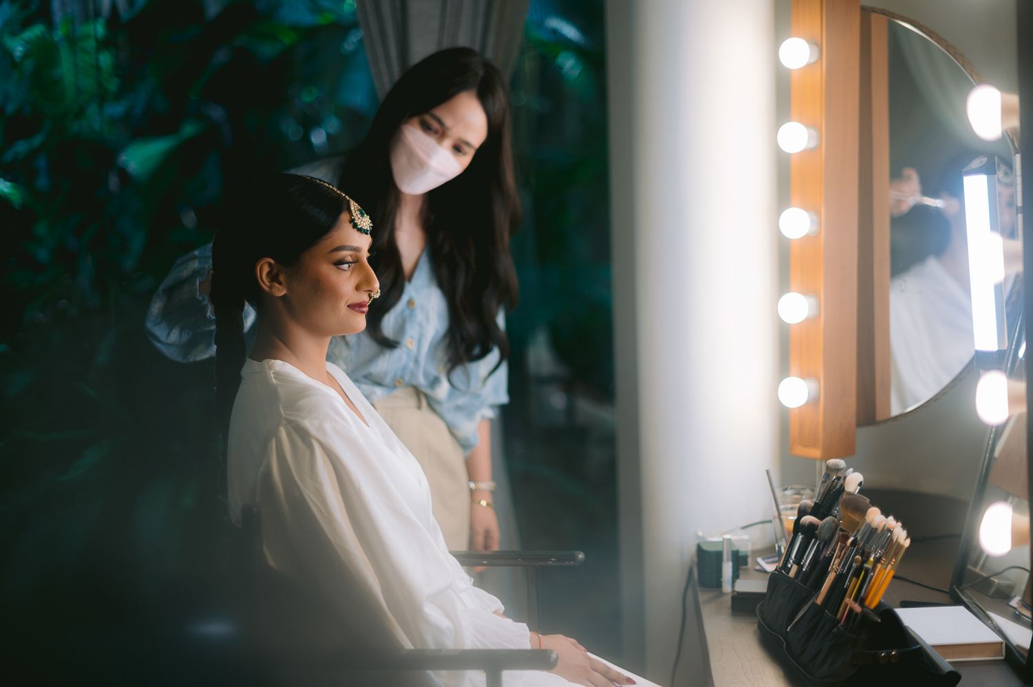 Makeup artist wearing a mask applies makeup to a seated woman in front of a lighted vanity mirror.