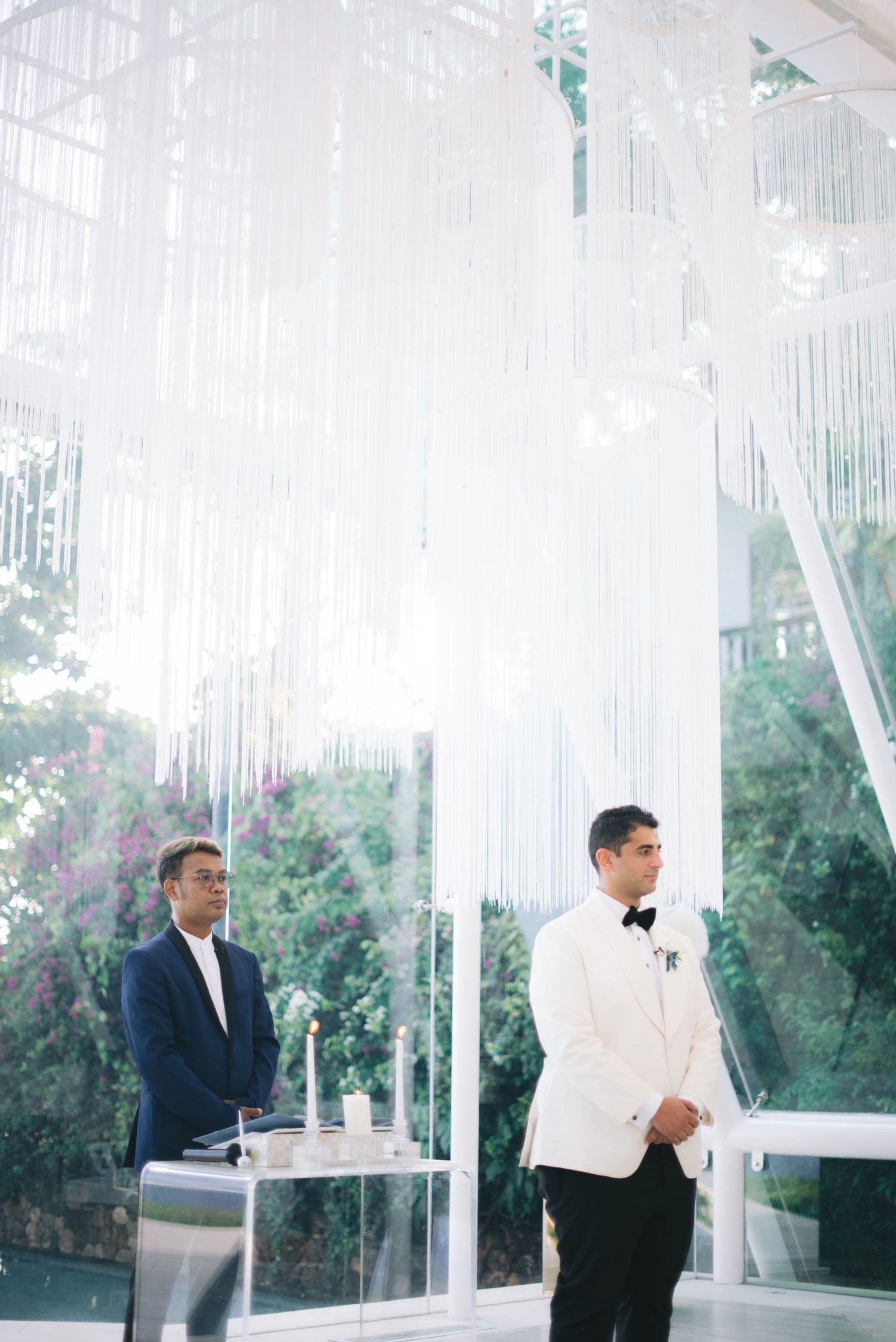 Groom in white tuxedo stands at outdoor wedding ceremony beneath elegant white fringe chandelier installation.