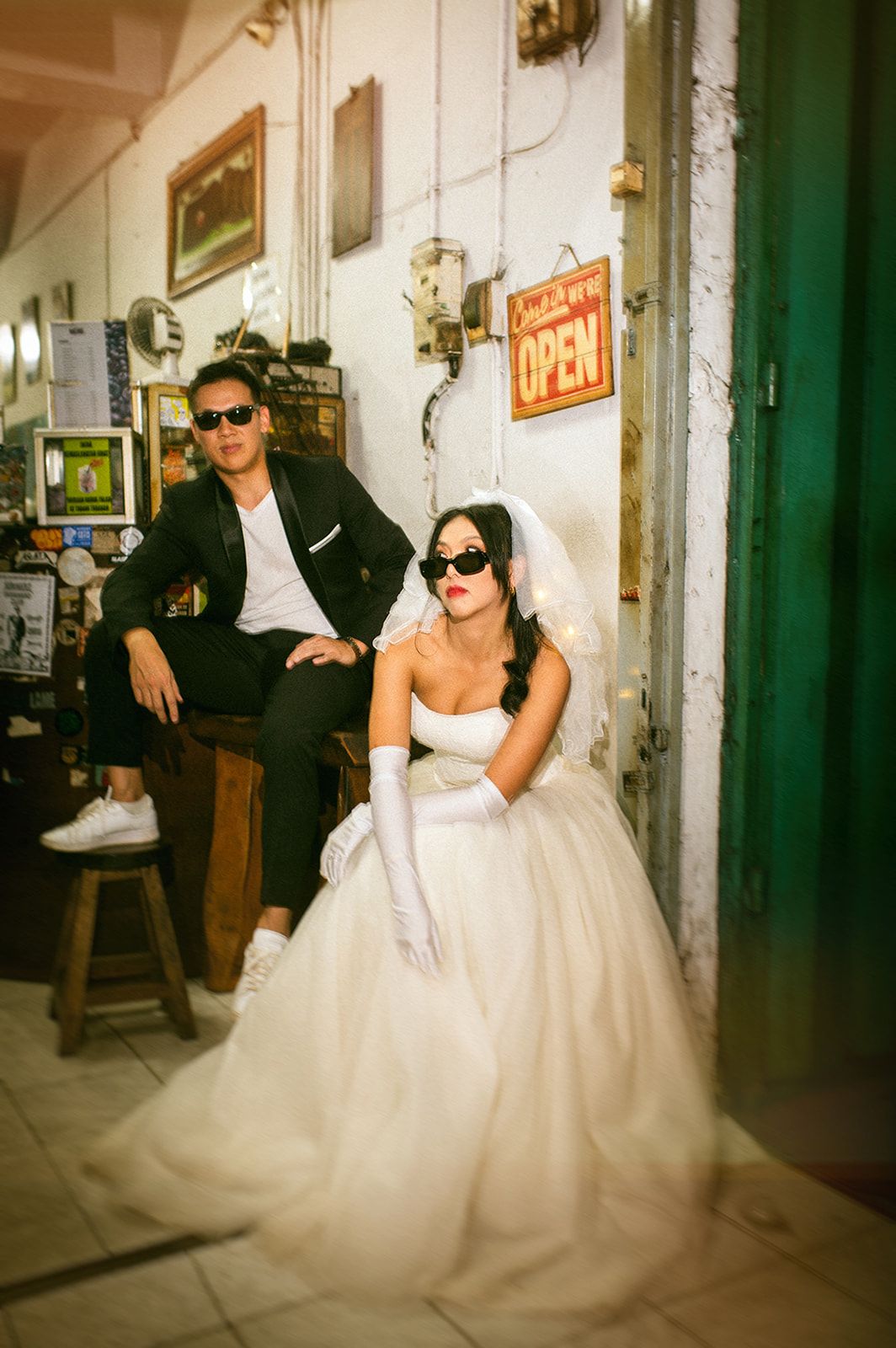 A bride in a white ballgown and gloves poses with a groom in a black suit, both wearing sunglasses in a vintage shop.