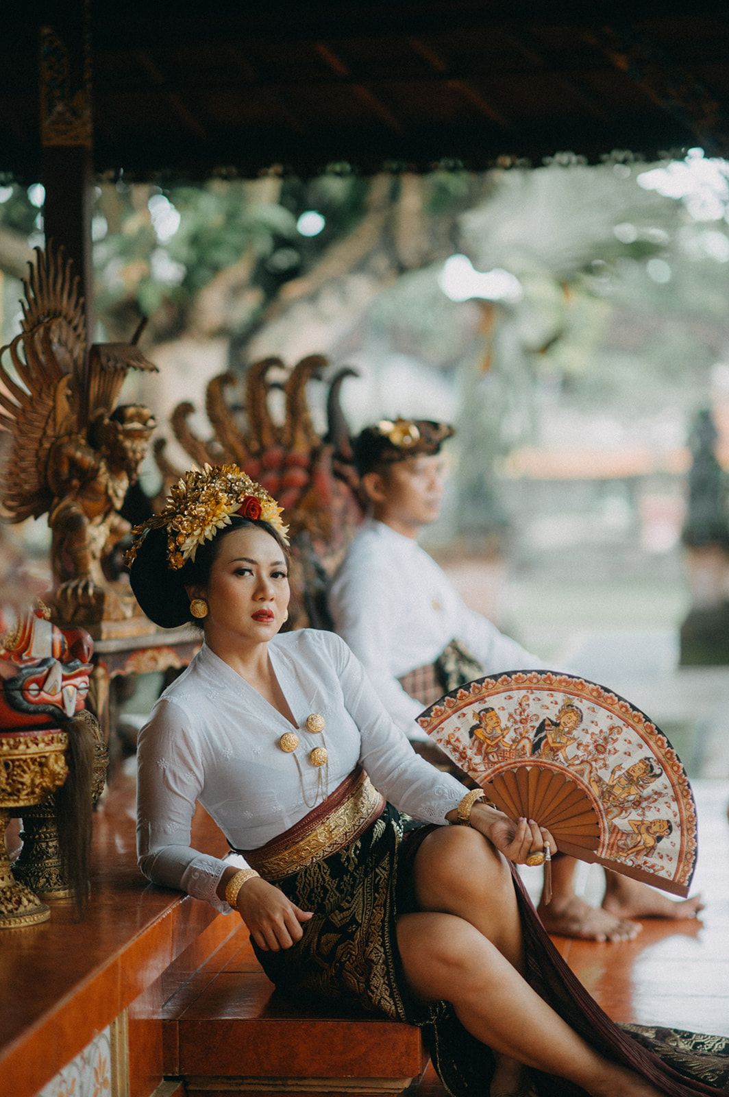 Two women in traditional Balinese attire with gold headdresses, one holding a decorative fan, seated at a temple.