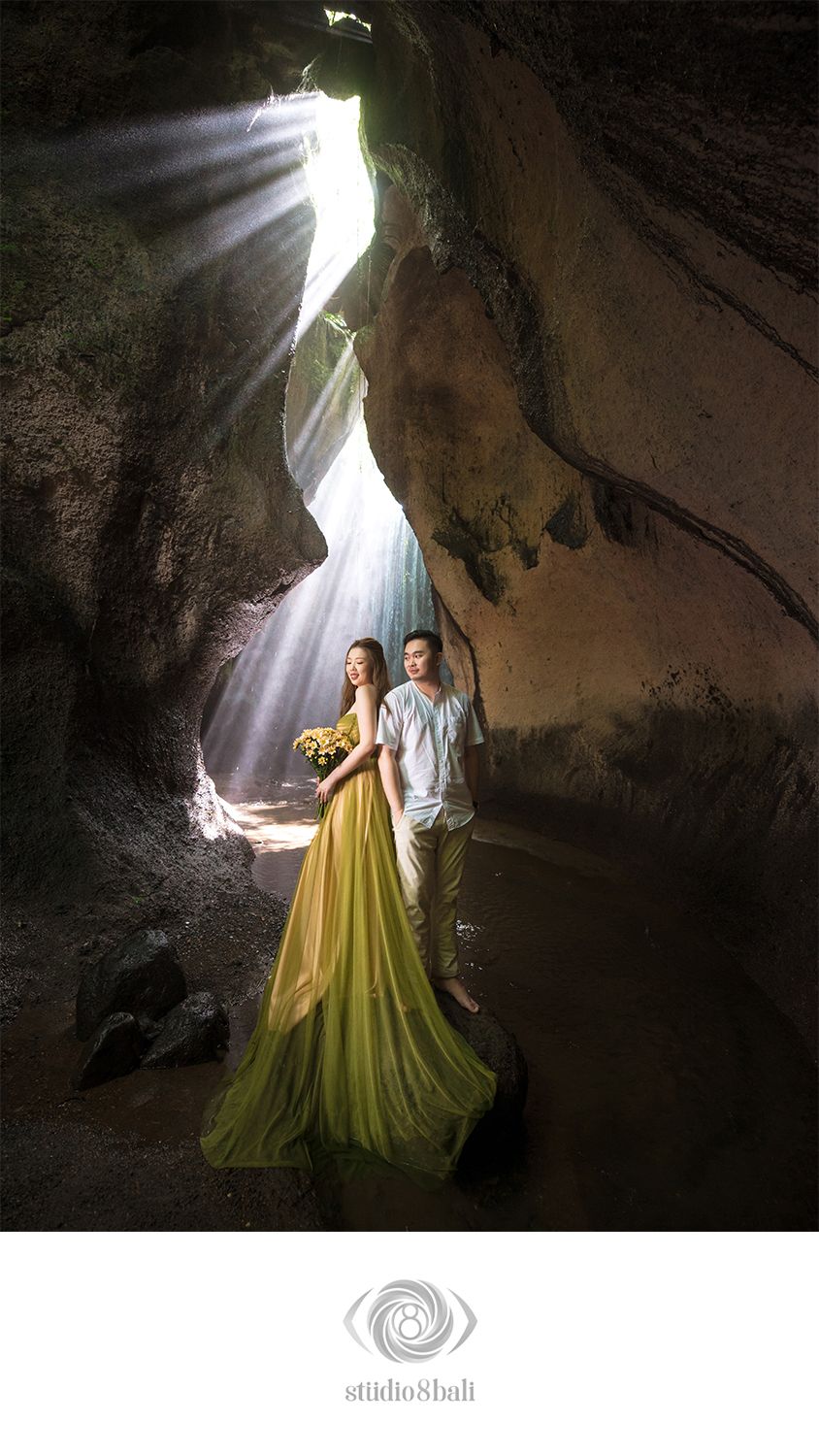 Couple in a cave with dramatic light rays, woman in green gown holding flowers, man in white shirt, Bali pre-wedding shoot.