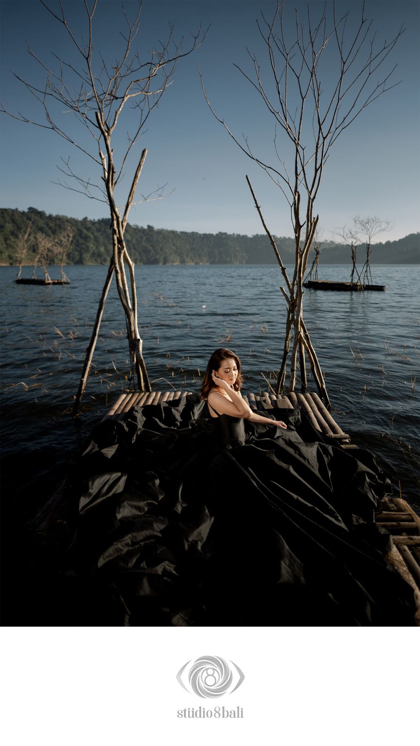 Woman in black dress on a bamboo raft surrounded by bare trees rising from a serene lake in Bali.