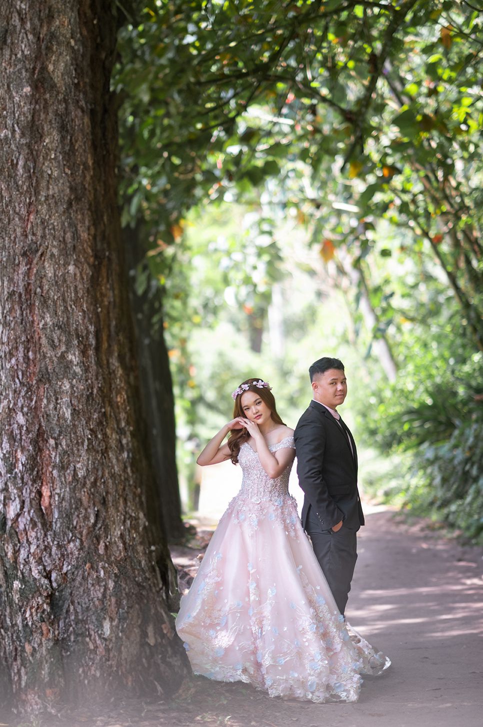 Couple in wedding attire posing under lush green tree canopy on a sunlit garden path.