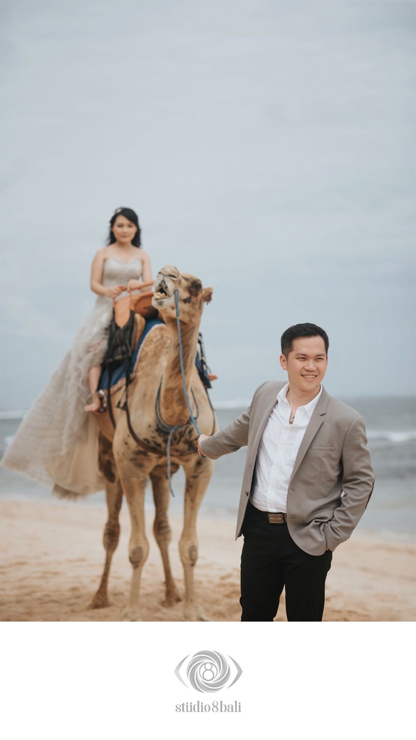 A groom leads a camel with his bride riding on the beach in an elegant pre-wedding photoshoot.
