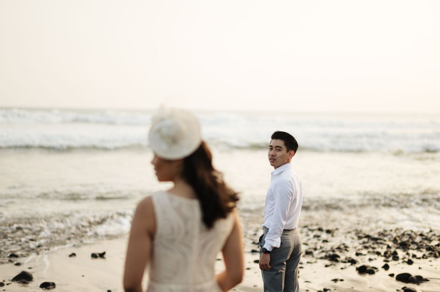 Couple in wedding attire on a beach at sunset, man in white shirt smiling back at woman in white dress and hat.