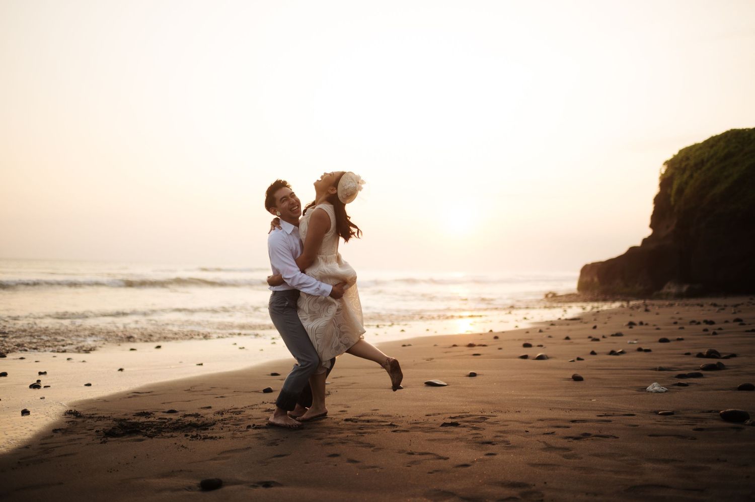Couple dancing joyfully on a sandy beach at sunset with ocean waves and rocky cliffs in the background.