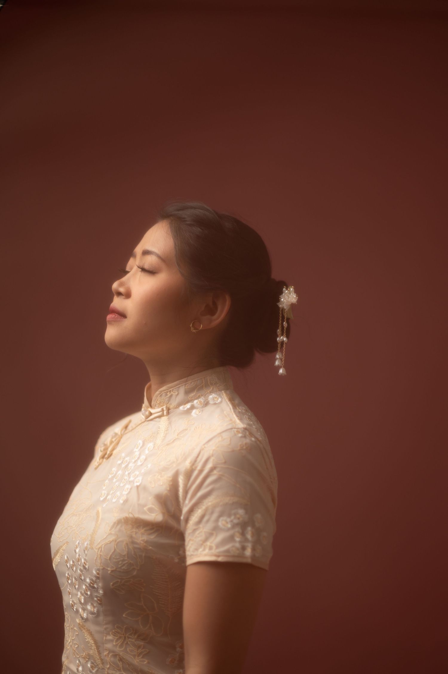 A young Asian woman in a white lace qipao with floral hair accessories poses gracefully with eyes closed against a red backdrop.