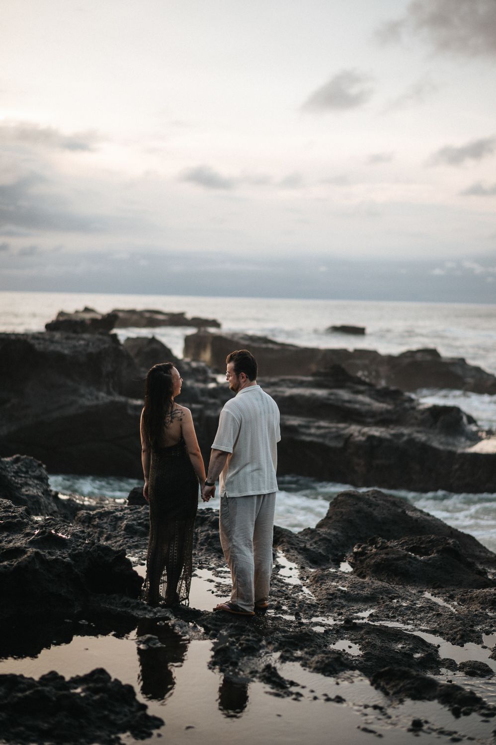 Couple holding hands on rocky ocean shore at dusk, waves and cloudy sky in background.