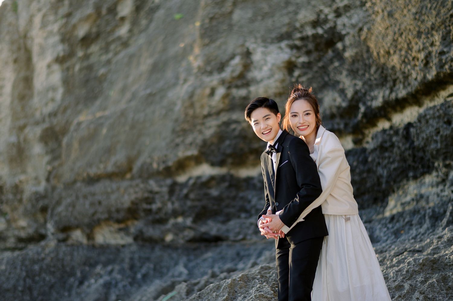 Happy Asian couple in wedding attire posing together against rocky cliffs for engagement photos.