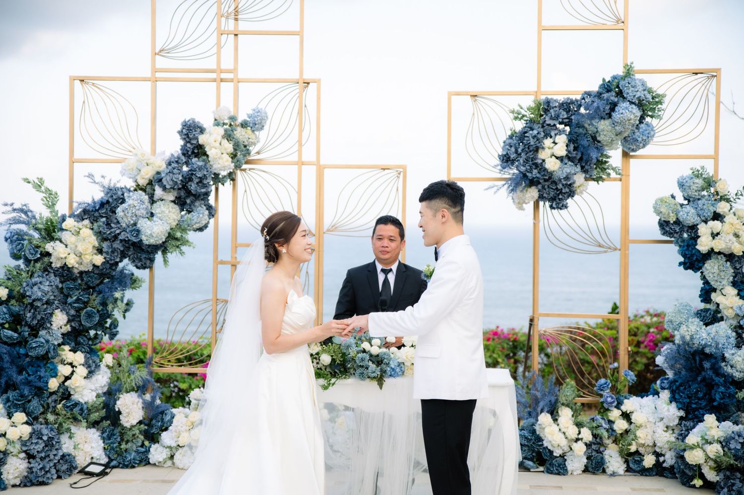 Bride in a white gown and groom in a white jacket exchange vows during a seaside wedding ceremony beneath blue and white floral arches.