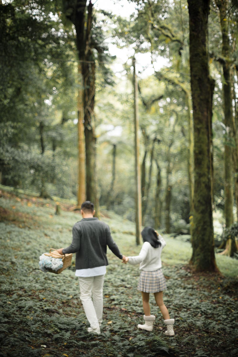 A couple walks hand-in-hand through a lush green forest, man carrying a picnic basket, woman in plaid skirt and boots.