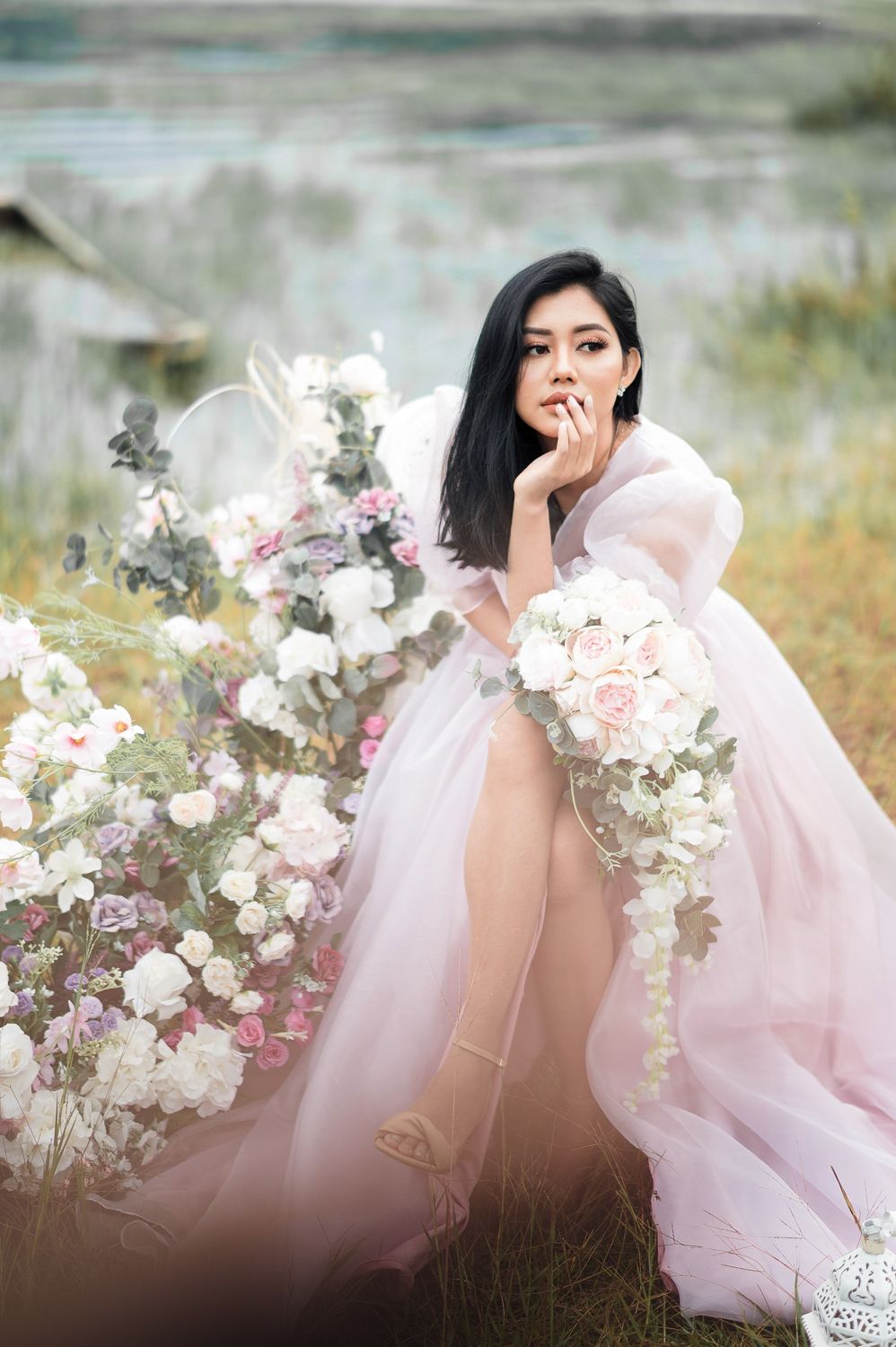 A bride in a pink tulle gown holds a white floral bouquet surrounded by blooms in a dreamy outdoor setting.