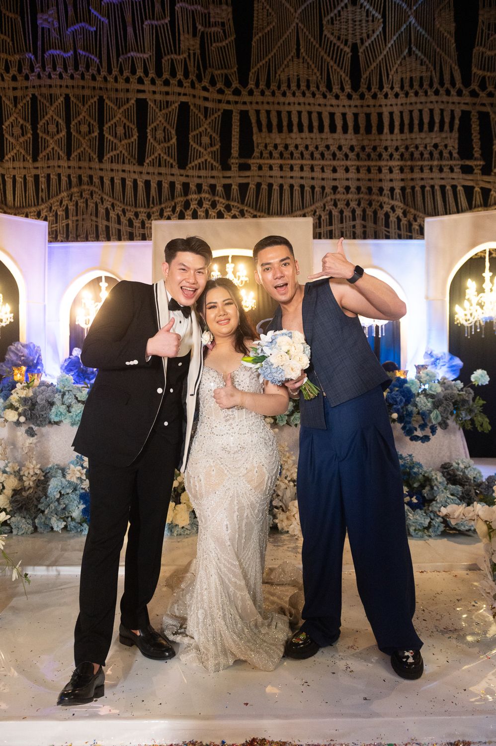 Bride in sequined gown poses with two men in formal attire at elegant wedding venue with blue floral decor.