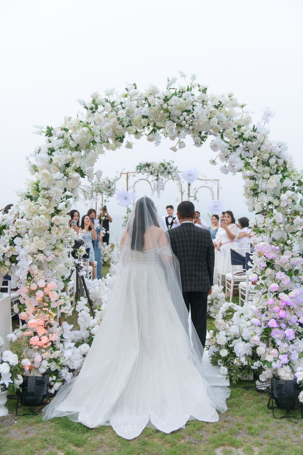 Bride in white gown and veil walks through floral arch adorned with white and pink flowers at wedding.