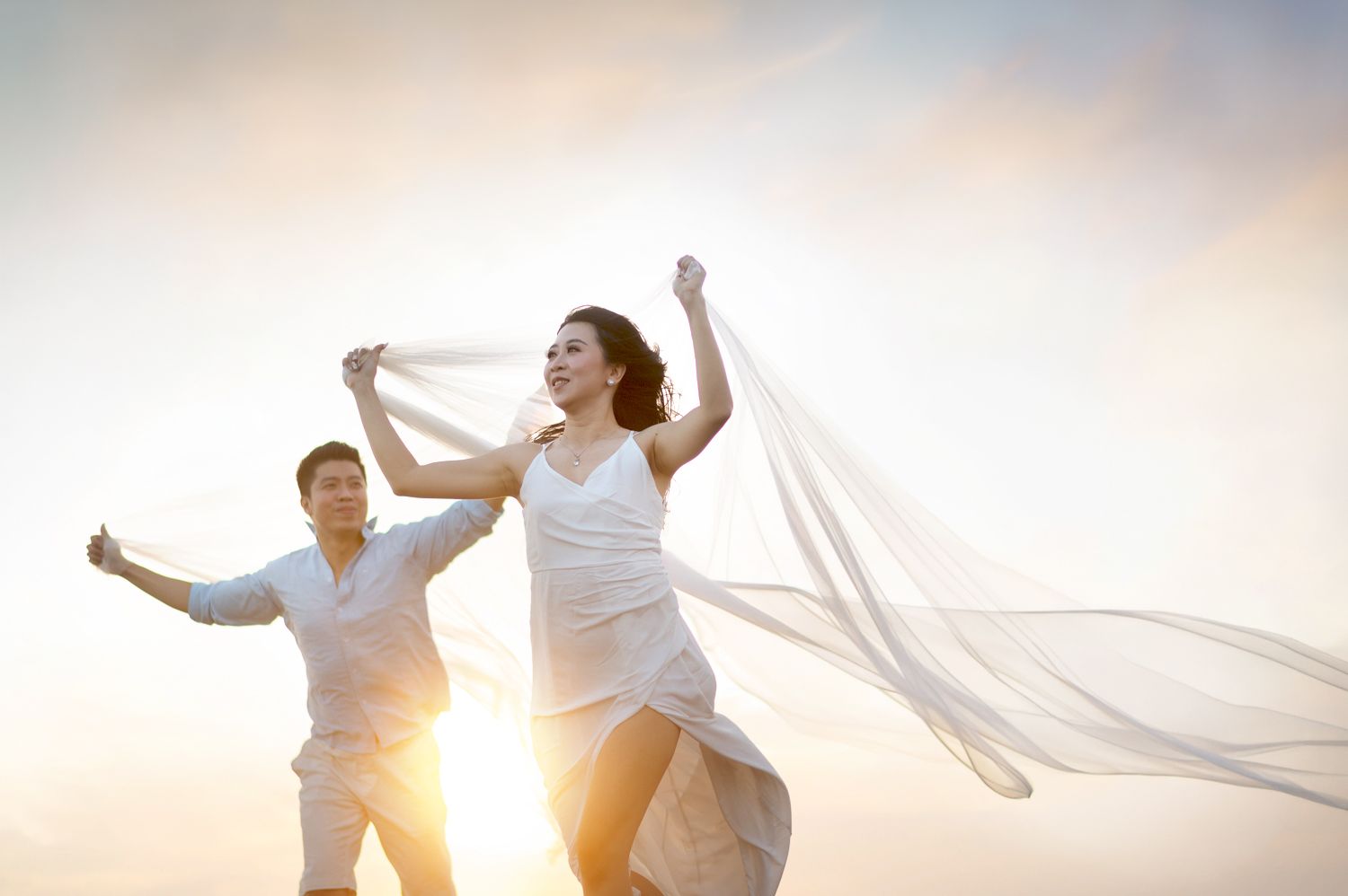 Joyful couple in white outfits running at sunset, woman's veil flowing in the wind against a golden sky.